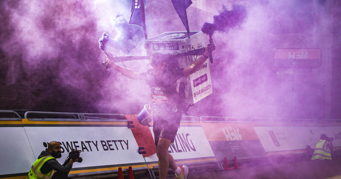 An image of The Man With a Fridge On His Back finishing the race, enveloped in purple smoke/fog. A fellow photographer looks on